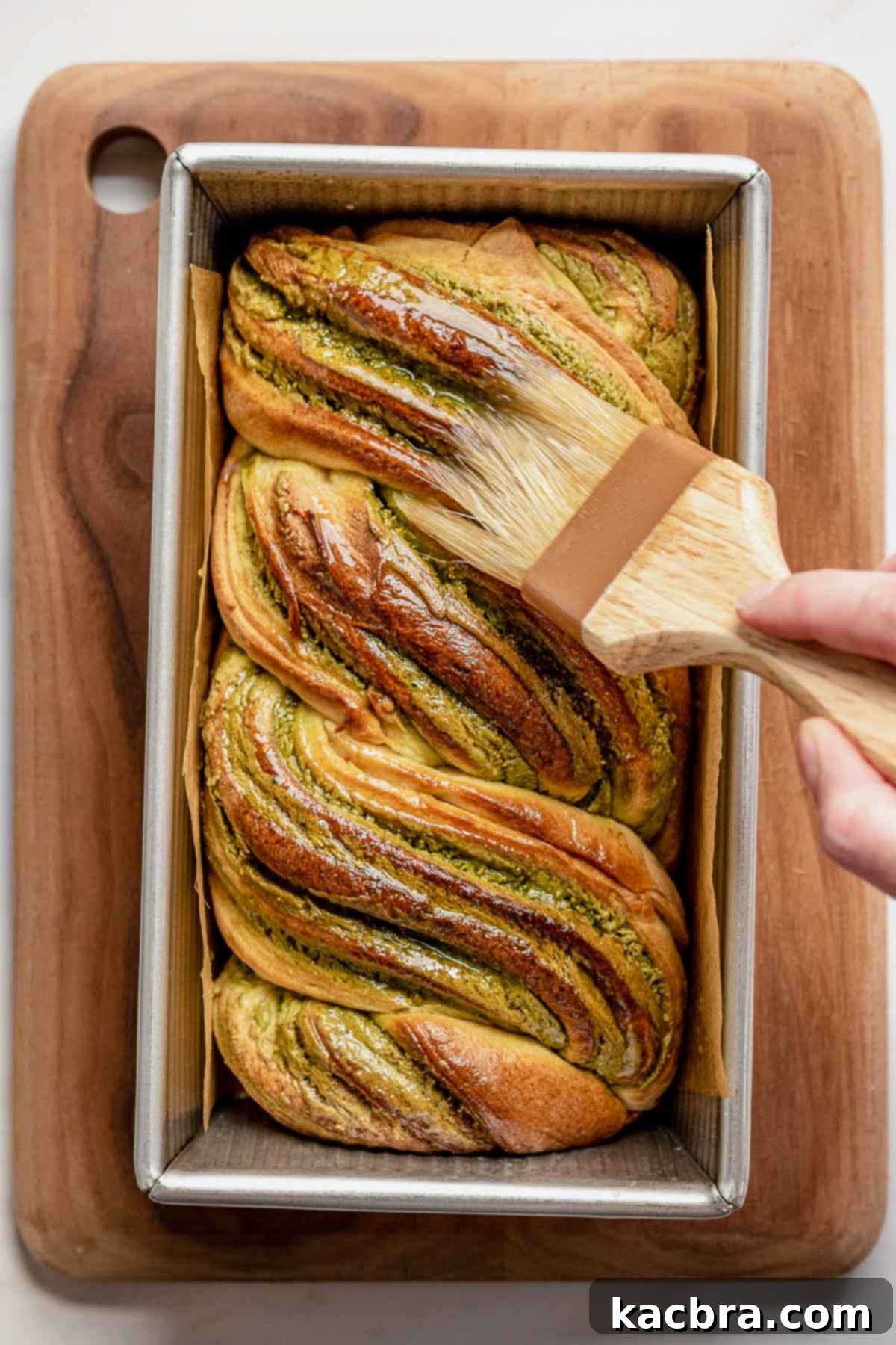A hand uses a pastry brush to apply a glistening layer of simple syrup over the top of the freshly baked, hot babka loaf, ensuring moisture and shine.