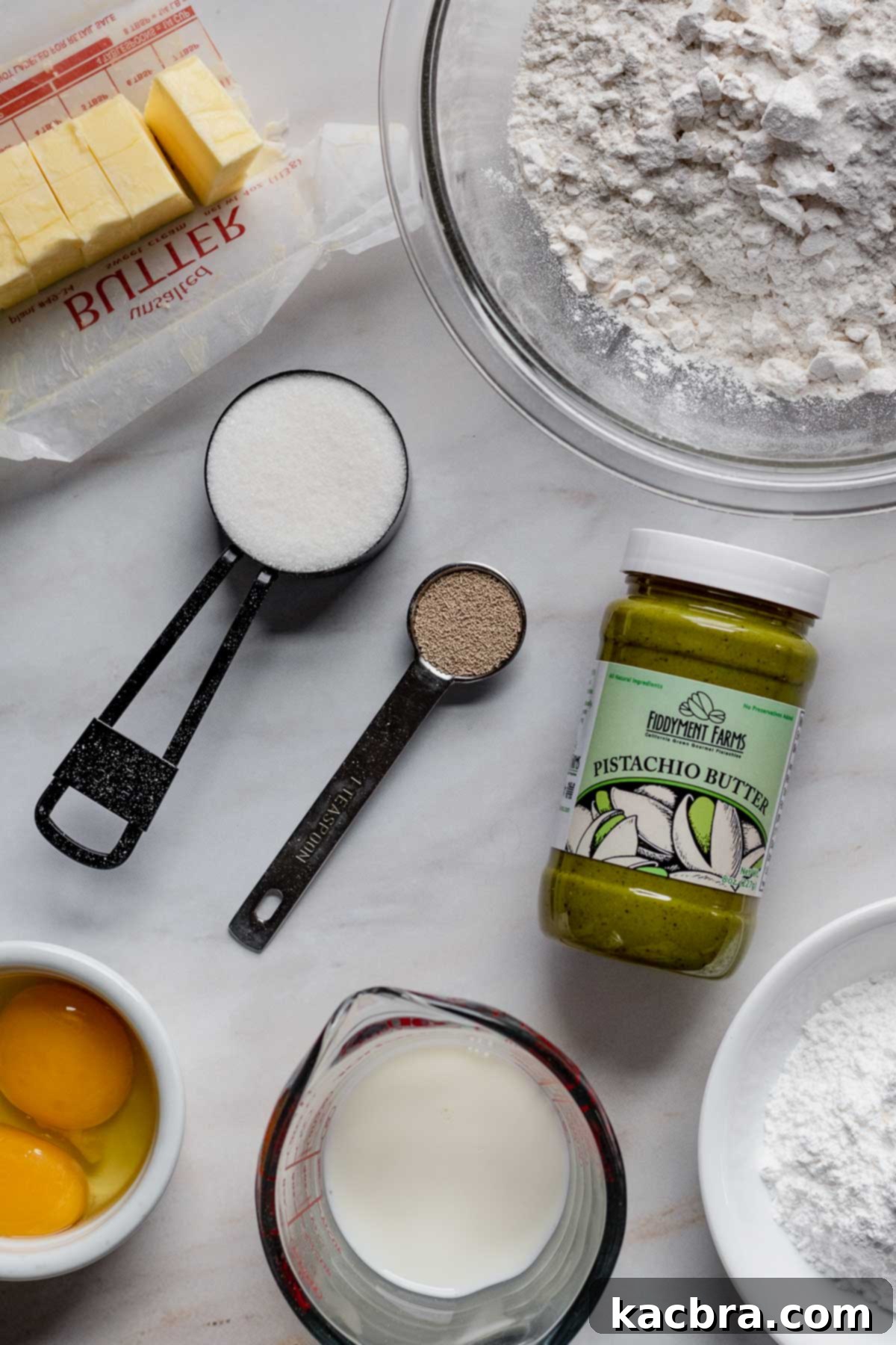 Assortment of fresh ingredients for pistachio babka, including butter, flour, sugar, active dry yeast, smooth pistachio butter, eggs, milk, and powdered sugar, neatly arranged on a rustic wooden table.