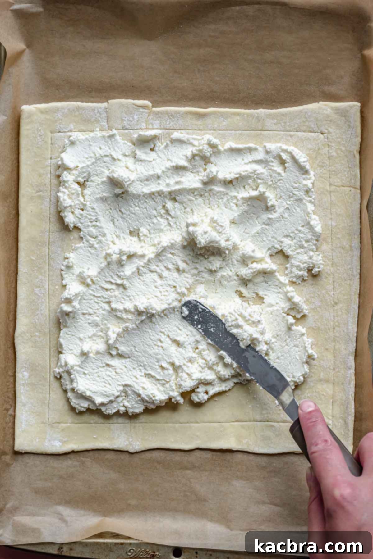 Rustic Tomato and Double Cheese Puff Pastry Tart 5 A spatula spreads cheese mixture onto puff pastry on a baking sheet.