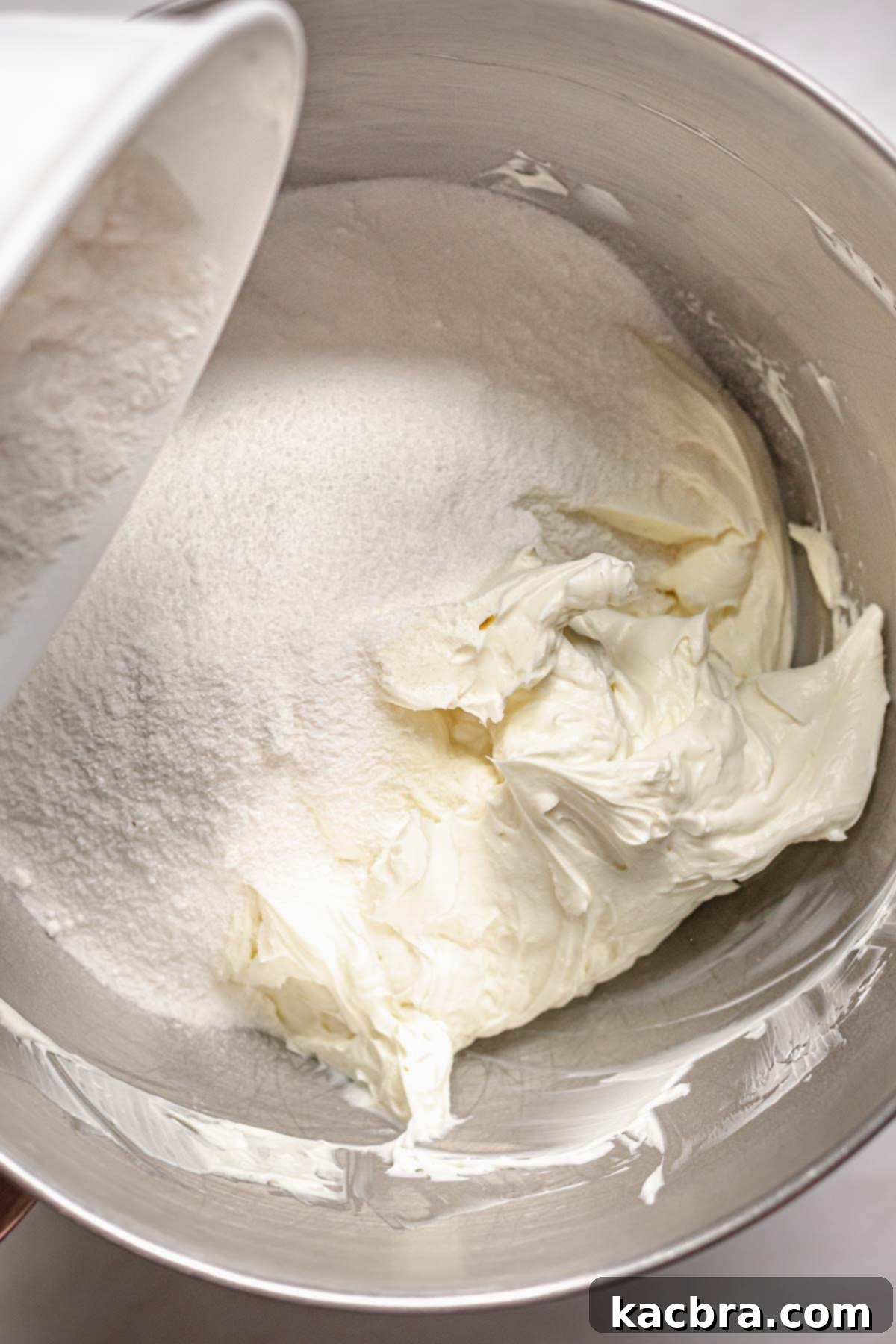 Adding granulated sugar to softened cream cheese in a mixing bowl for the cheesecake batter.