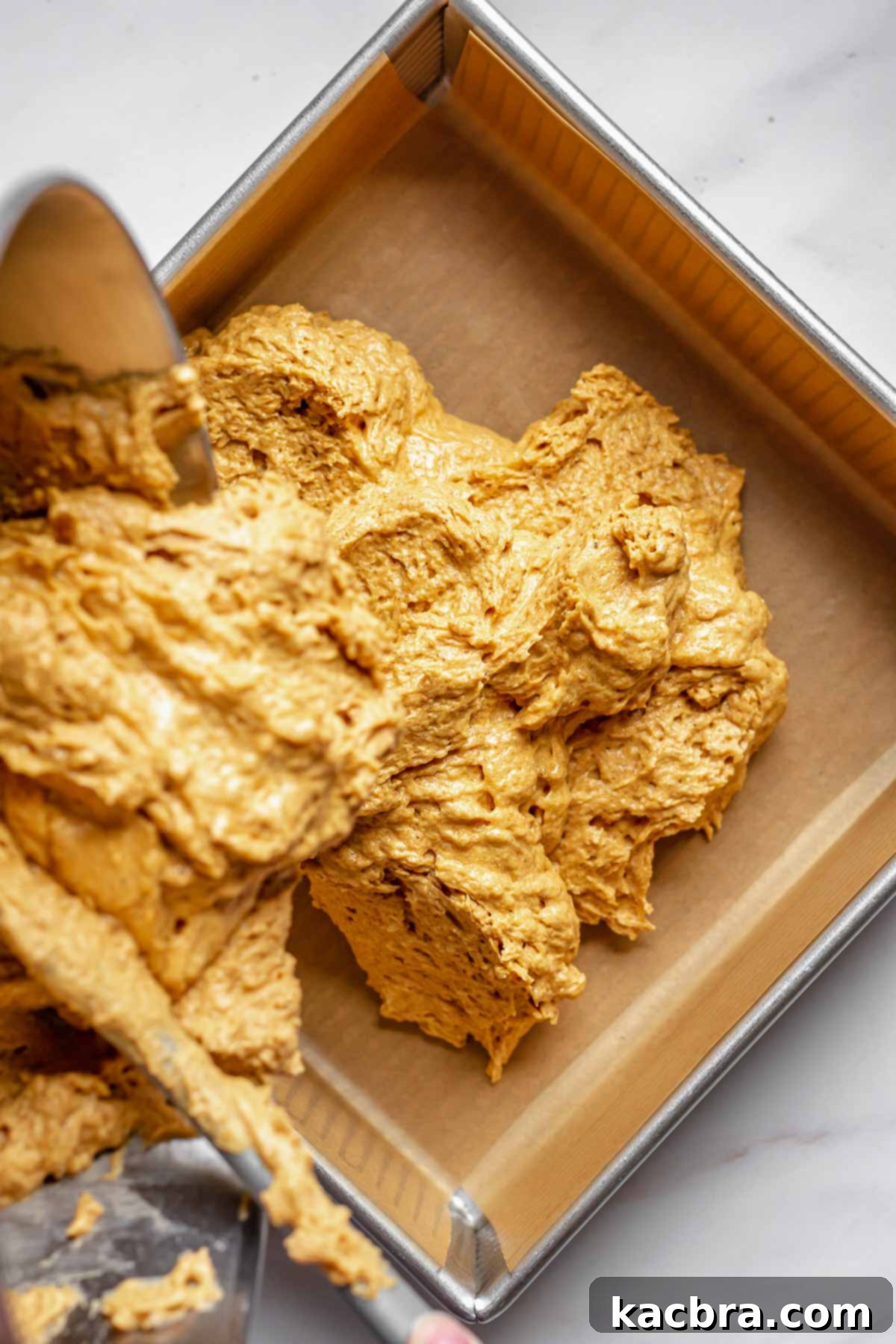 Smooth cake batter being poured into a prepared square baking pan lined with parchment paper.