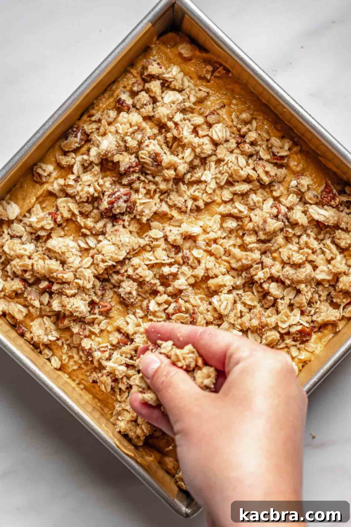 The pecan streusel topping being sprinkled evenly over the pumpkin cake batter in the pan.