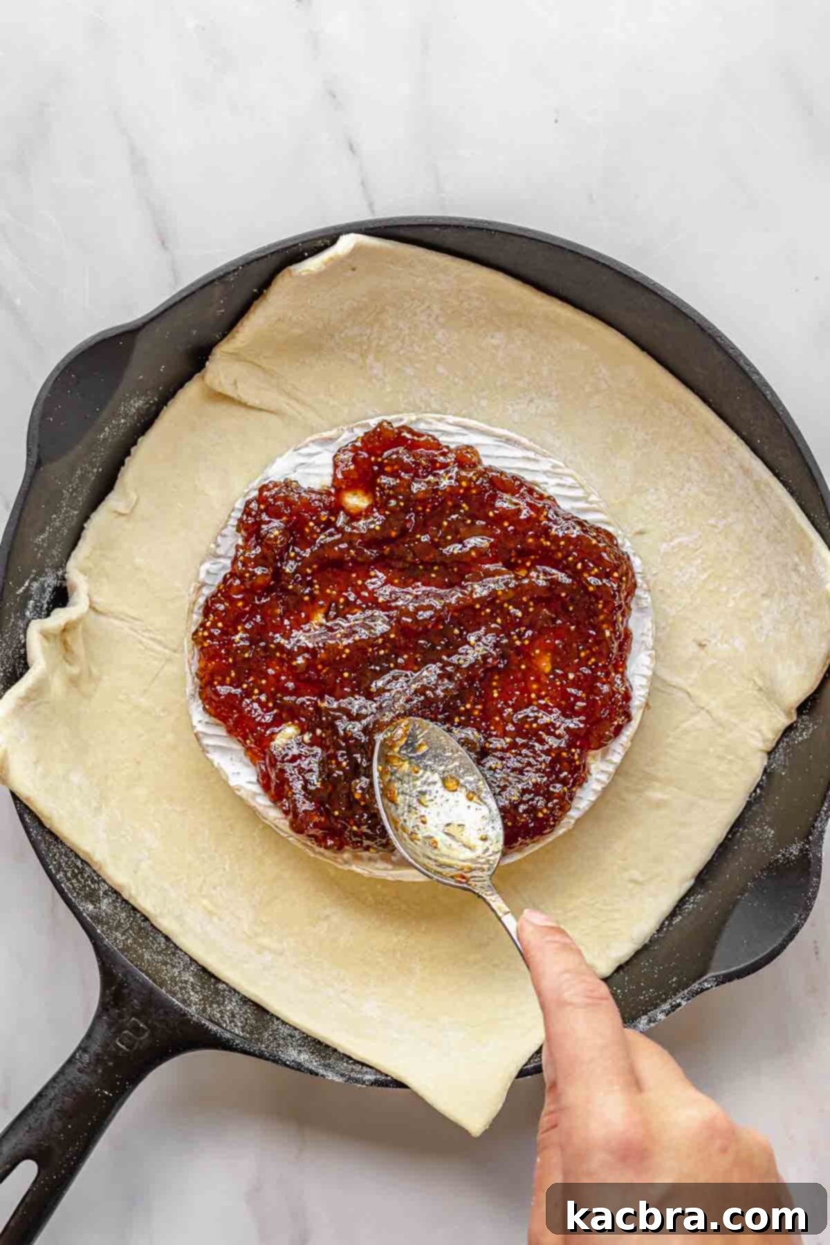 Fig jam being generously spread over the scored brie cheese with a spoon.