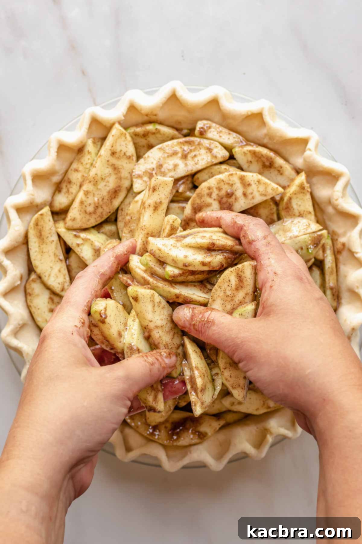 Hands adding spiced apple slices to a pie crust.