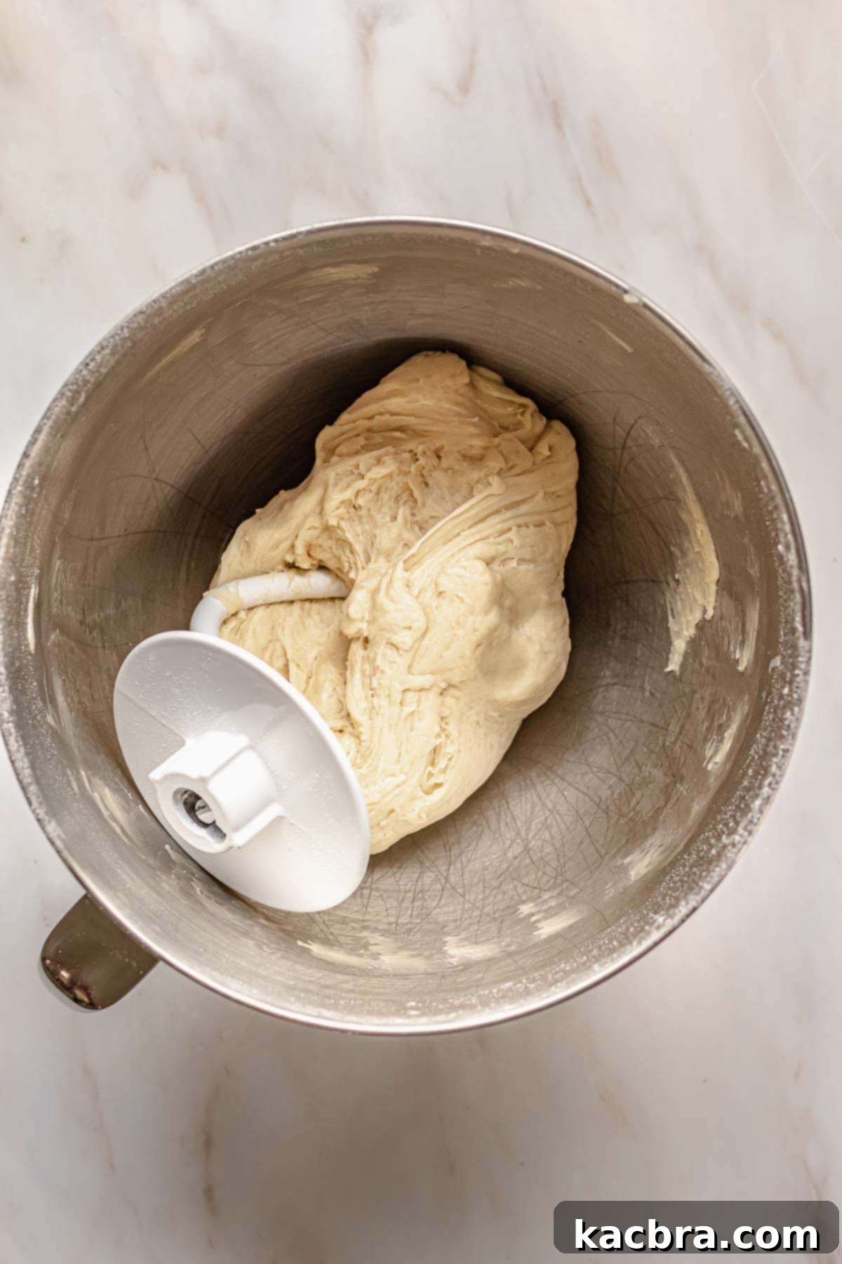 Smooth, elastic dough clinging to the dough hook in a stand mixer bowl after sufficient kneading.