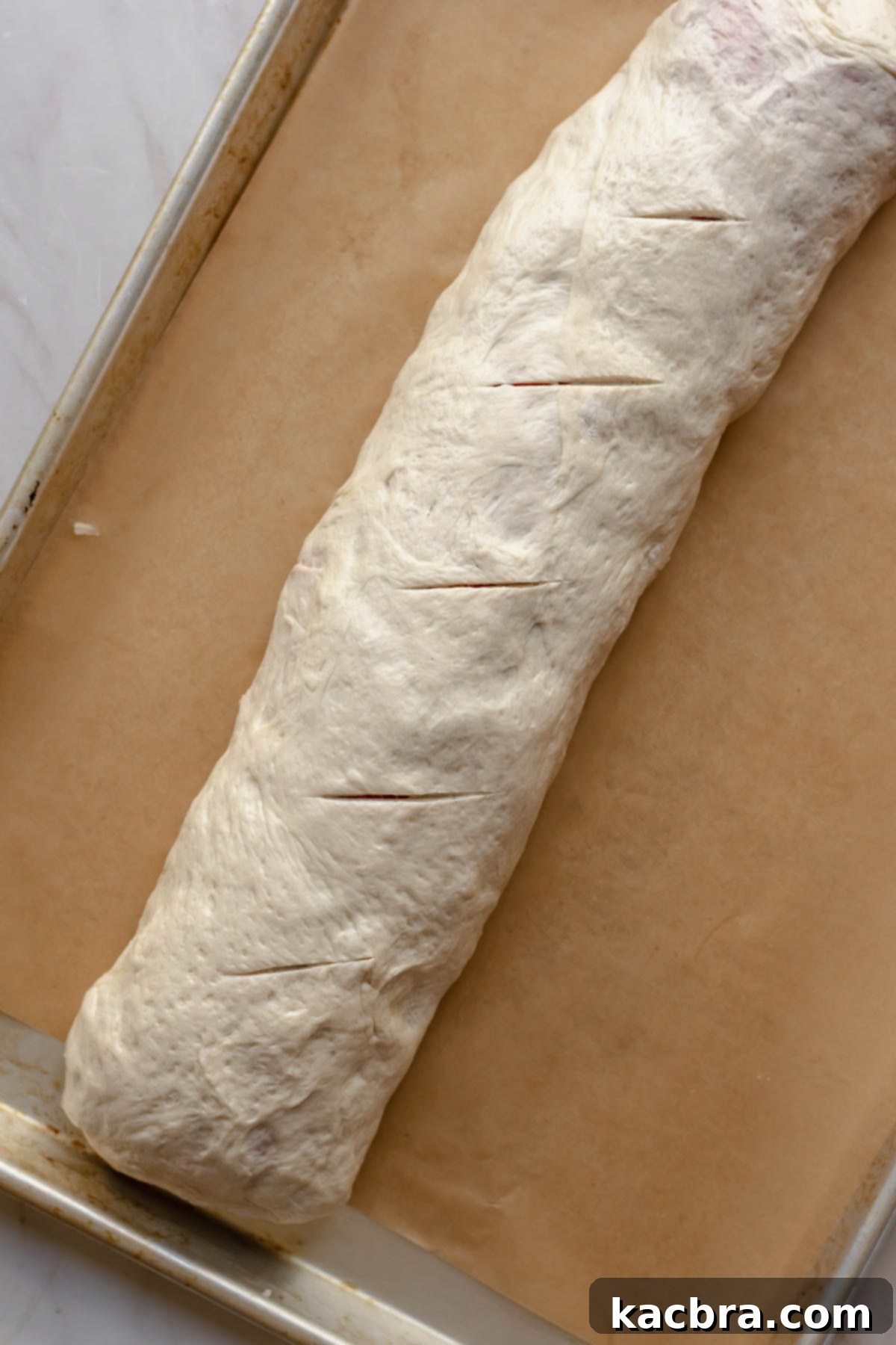 A log of pepperoni roll on a sheet pan with slits in the top.