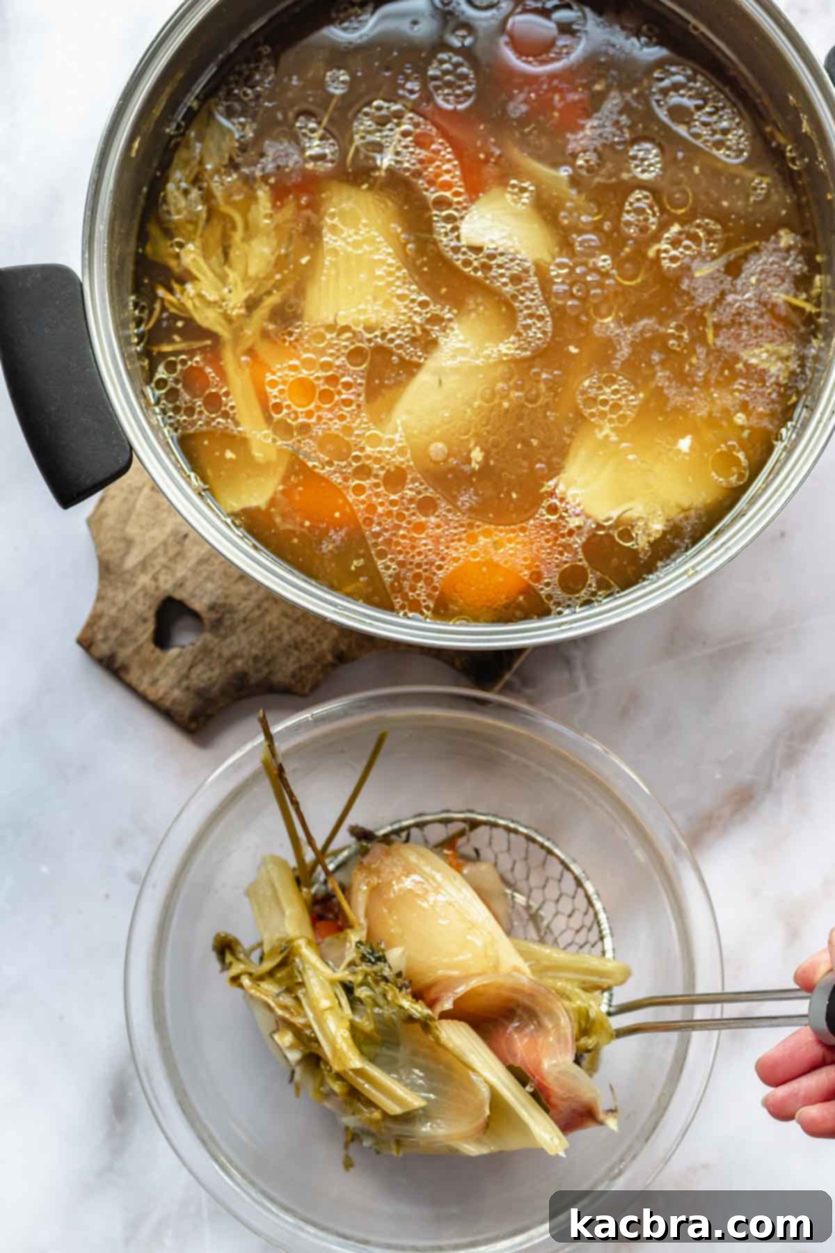 A hand uses a spider to remove cooked vegetables from chicken stock.