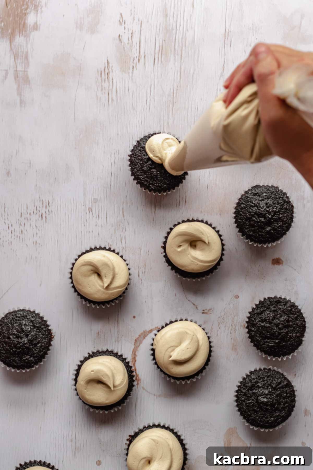 A hand expertly pipes swirls of creamy espresso buttercream onto cooled chocolate mocha cupcakes, preparing them for their final garnish.