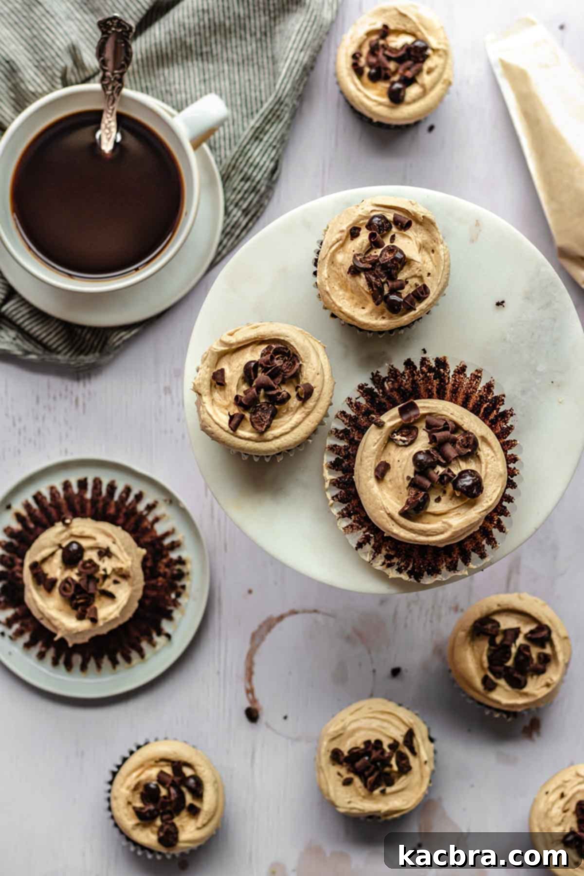 Finished mocha cupcakes are scattered on a counter, some adorned with chocolate curls and chocolate-covered coffee beans, with coffee beans thoughtfully placed alongside.