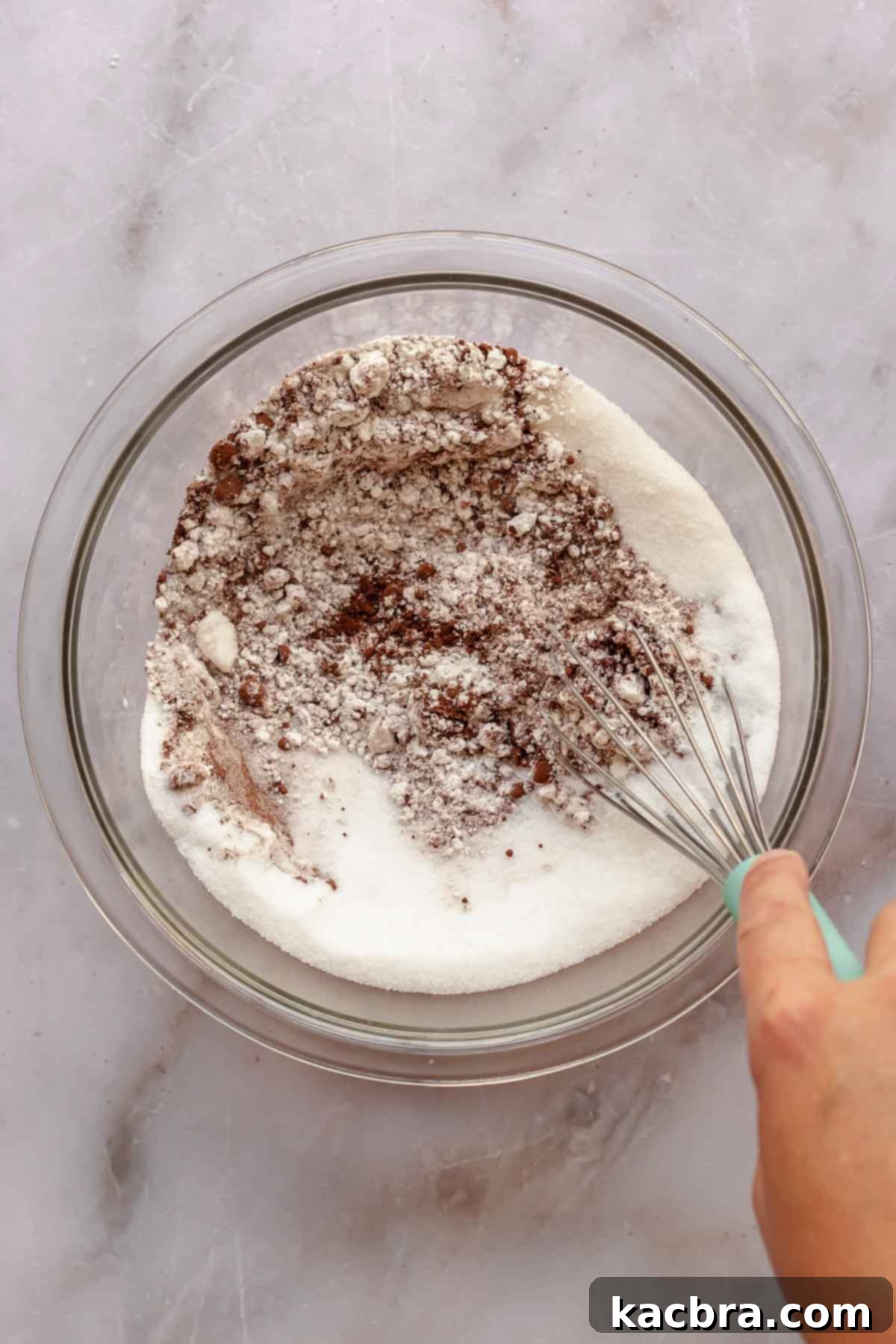 A hand holds a whisk, mixing together dry ingredients like flour, sugar, and cocoa powder in a large mixing bowl, creating the foundation for the cupcake batter.