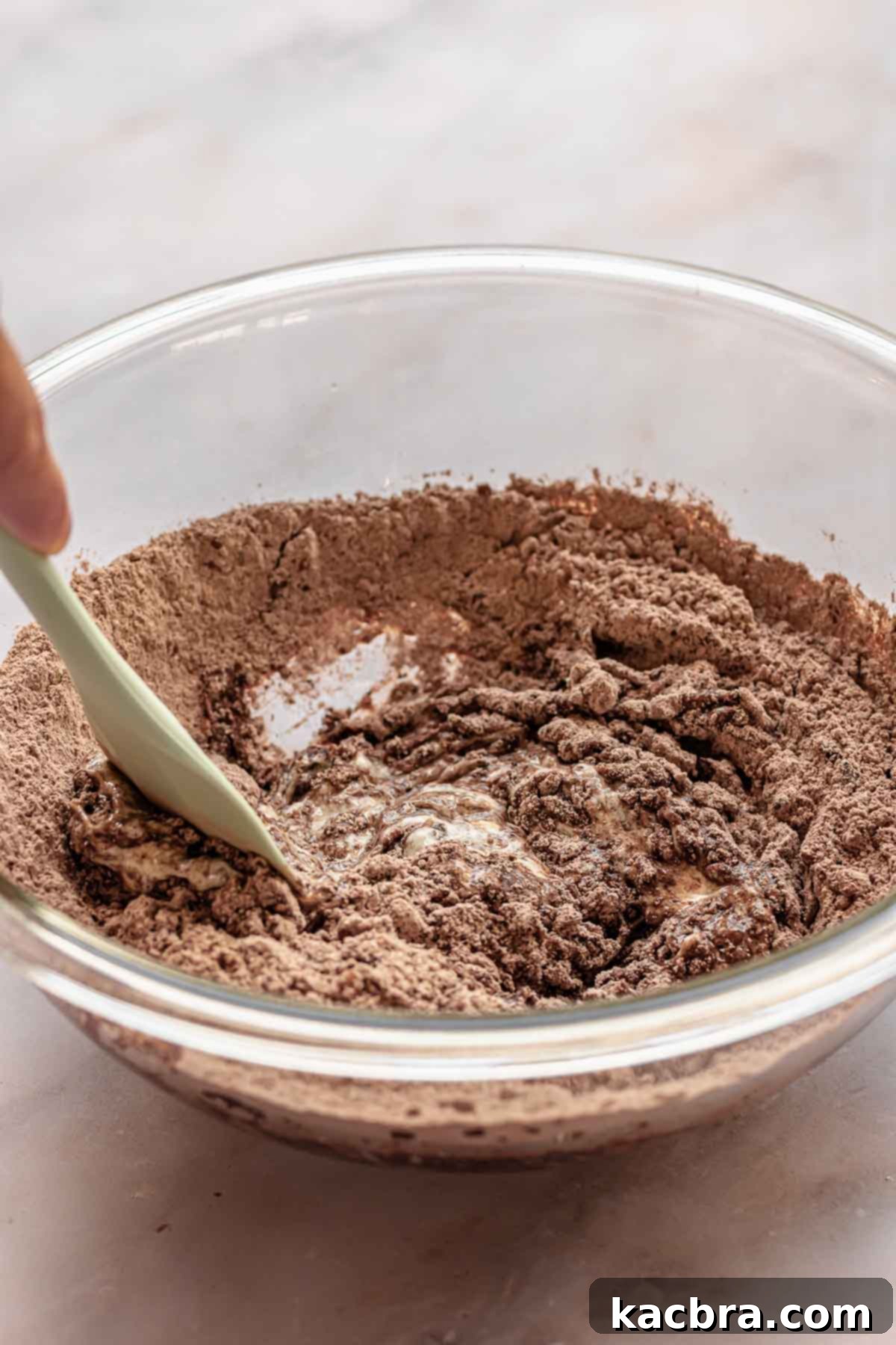 A hand gently folds the wet and dry cupcake ingredients together in a mixing bowl with a rubber spatula, gradually combining them into a cohesive batter.