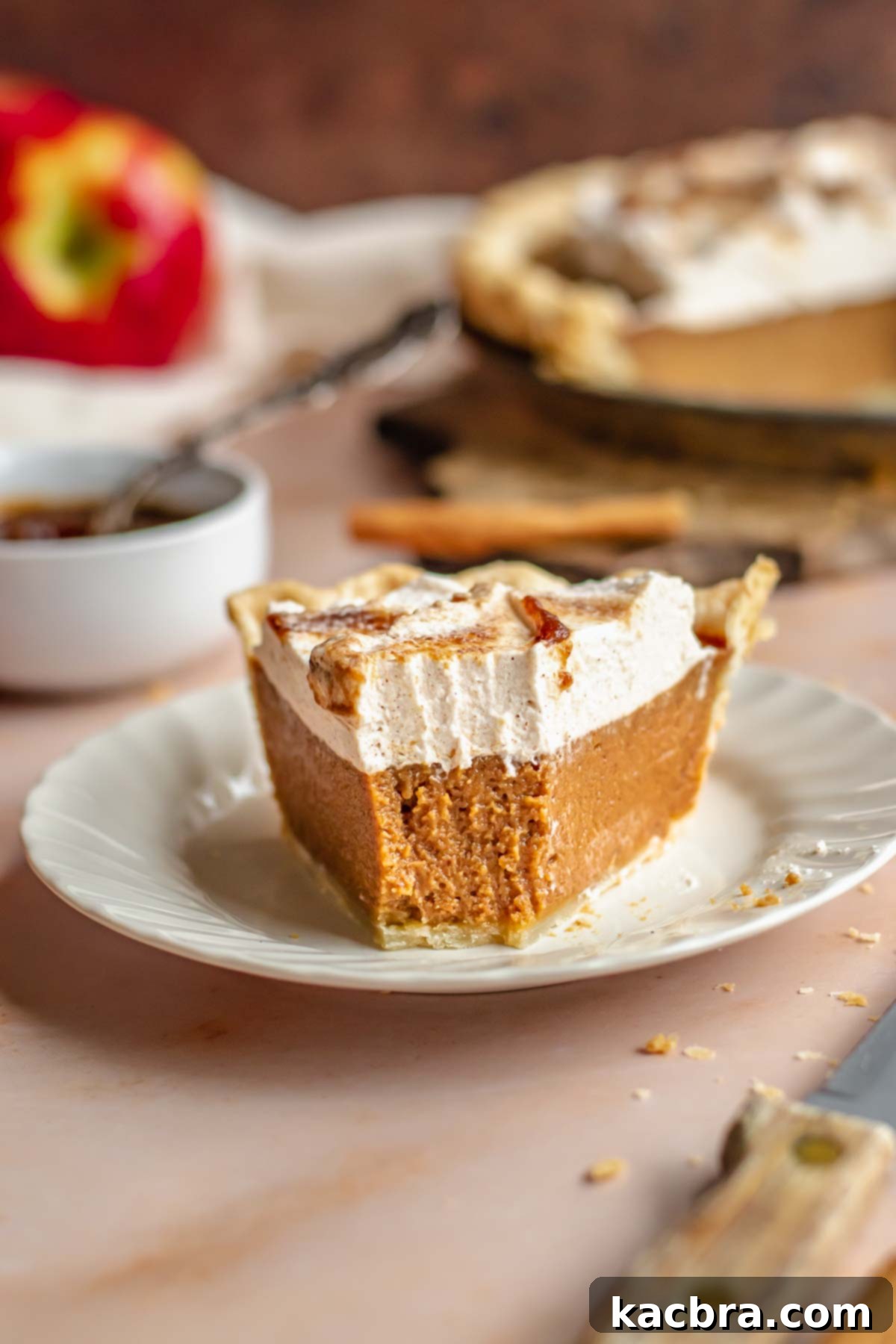 A perfectly sliced apple butter pie rests on a delicate white plate, with a small bite removed to showcase its smooth, rich custard filling and flaky crust. The focus is on the inviting texture and irresistible appearance of this fall-inspired dessert.