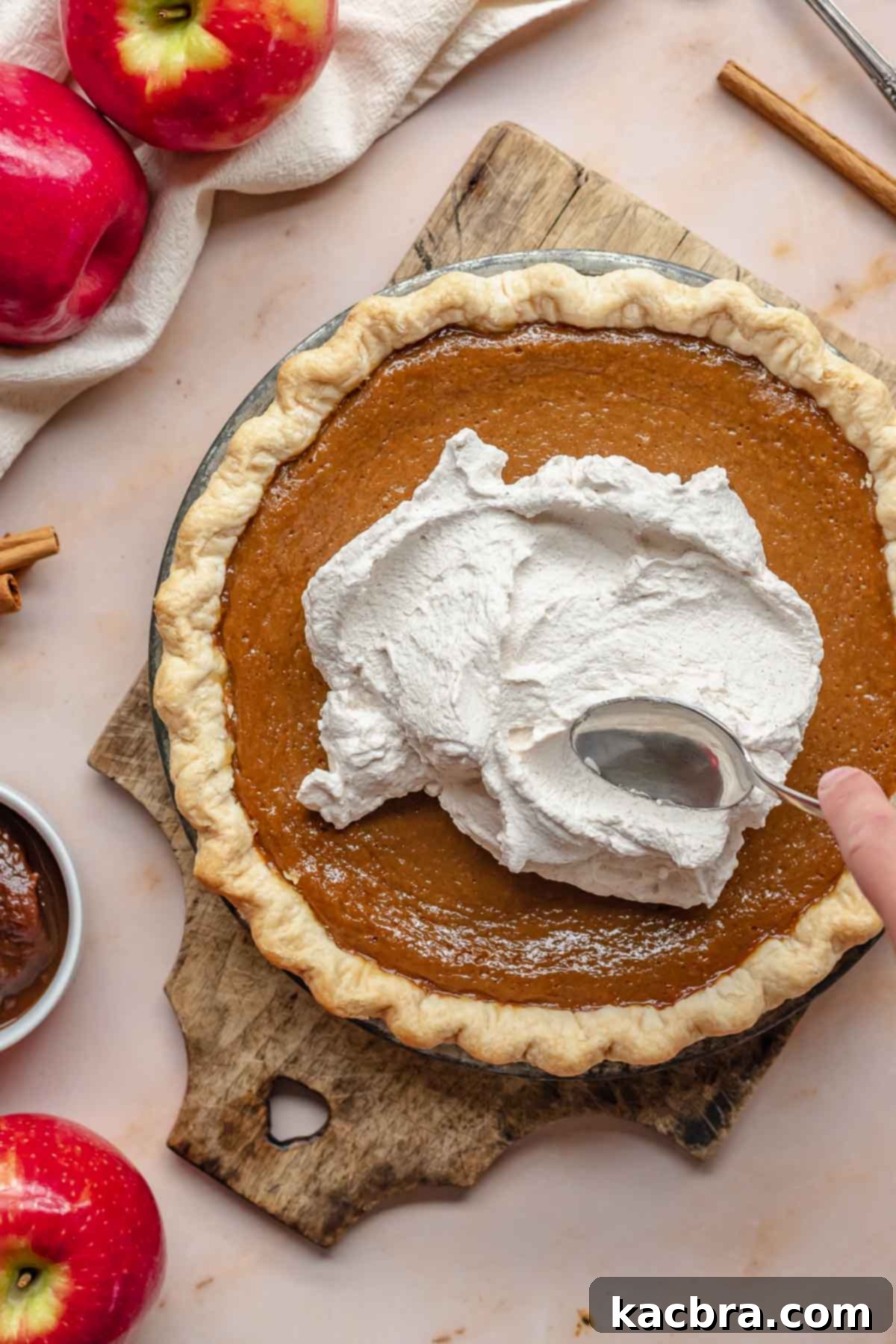 A spoon gracefully sweeps a generous dollop of cinnamon whipped cream onto a freshly baked apple butter pie, creating an inviting presentation.