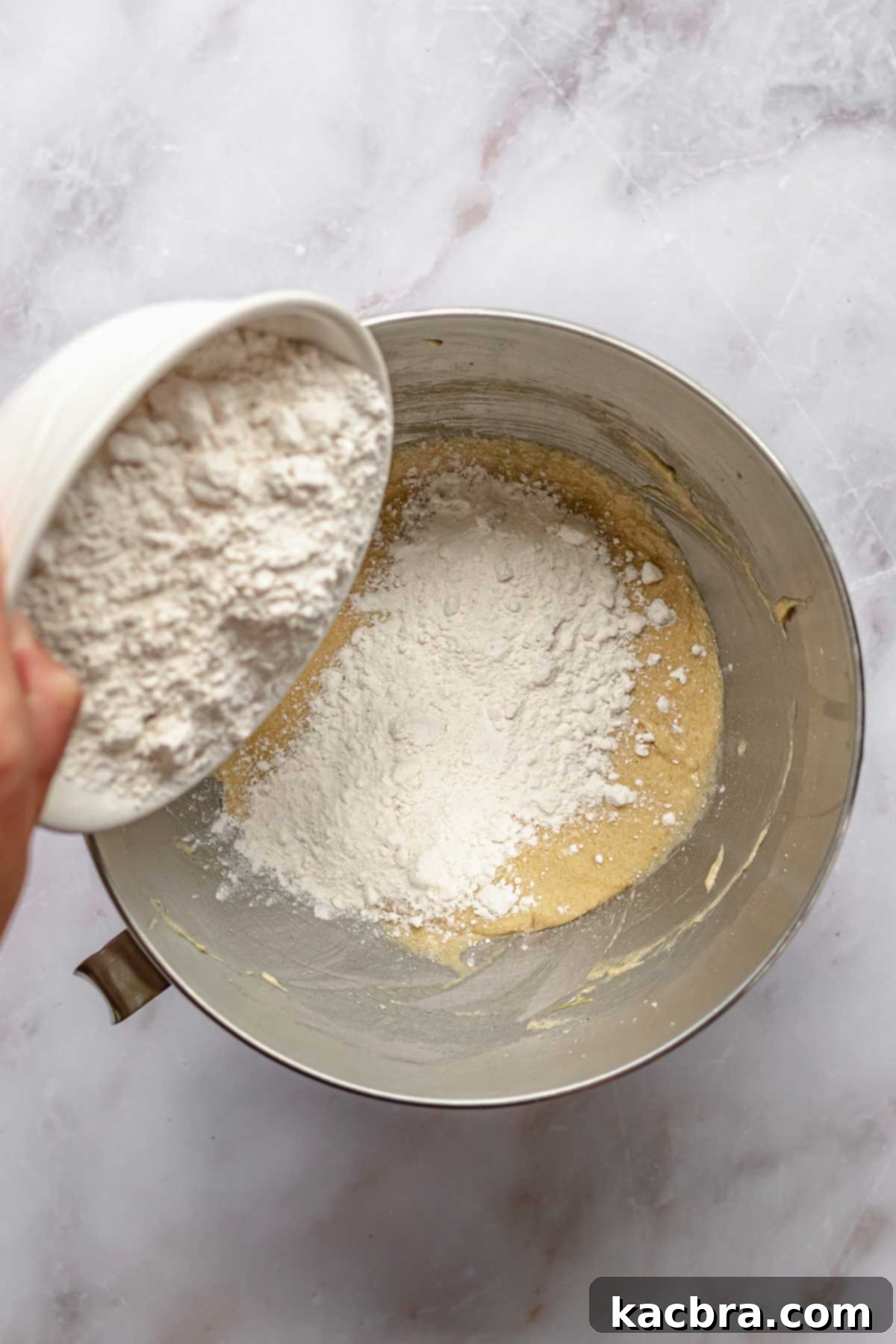 A hand pouring a bowl of dry cake ingredients into a stand mixer bowl containing wet ingredients.