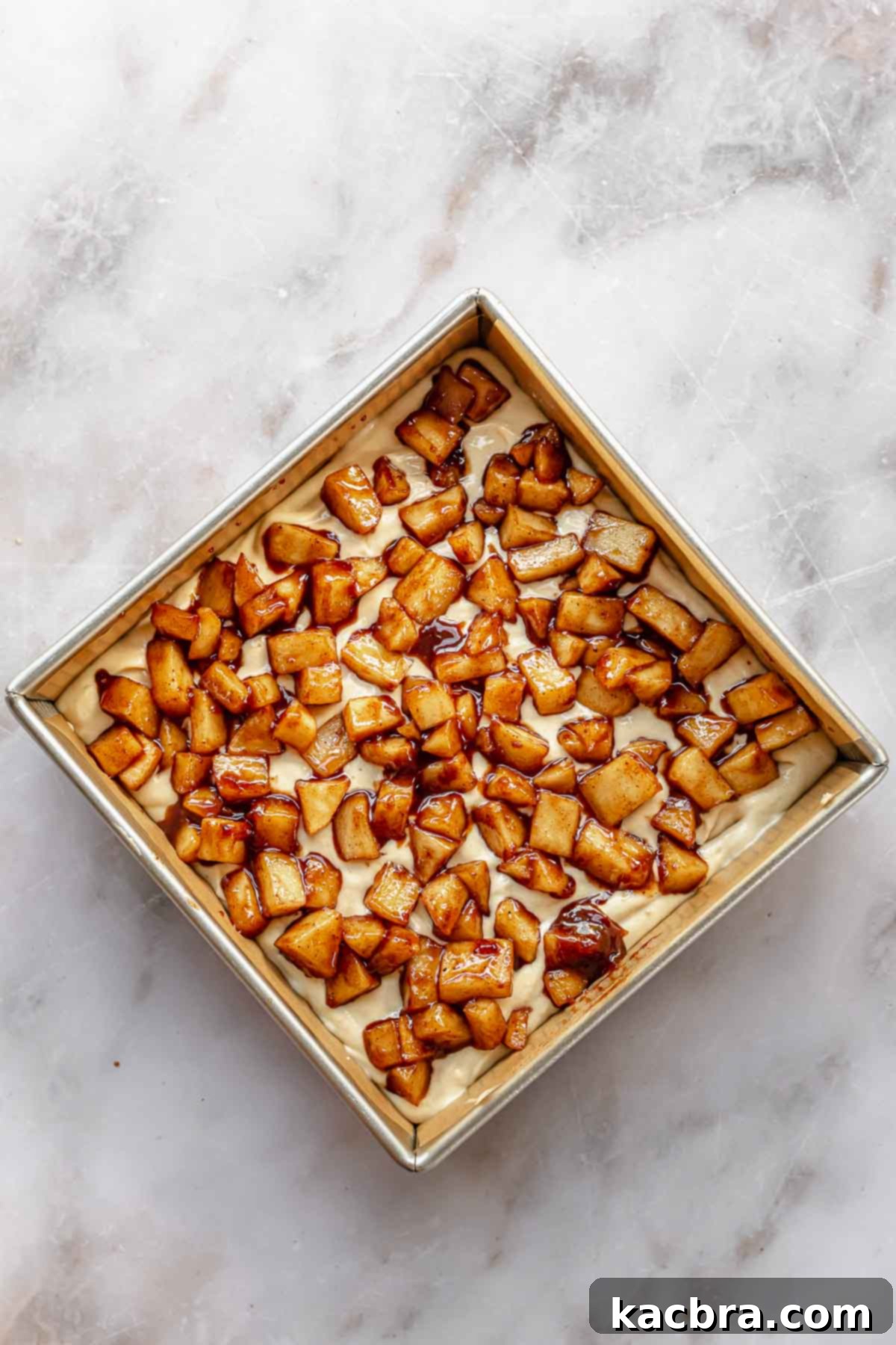 Cinnamon apple chunks spread evenly on top of raw cake batter in a square baking pan.