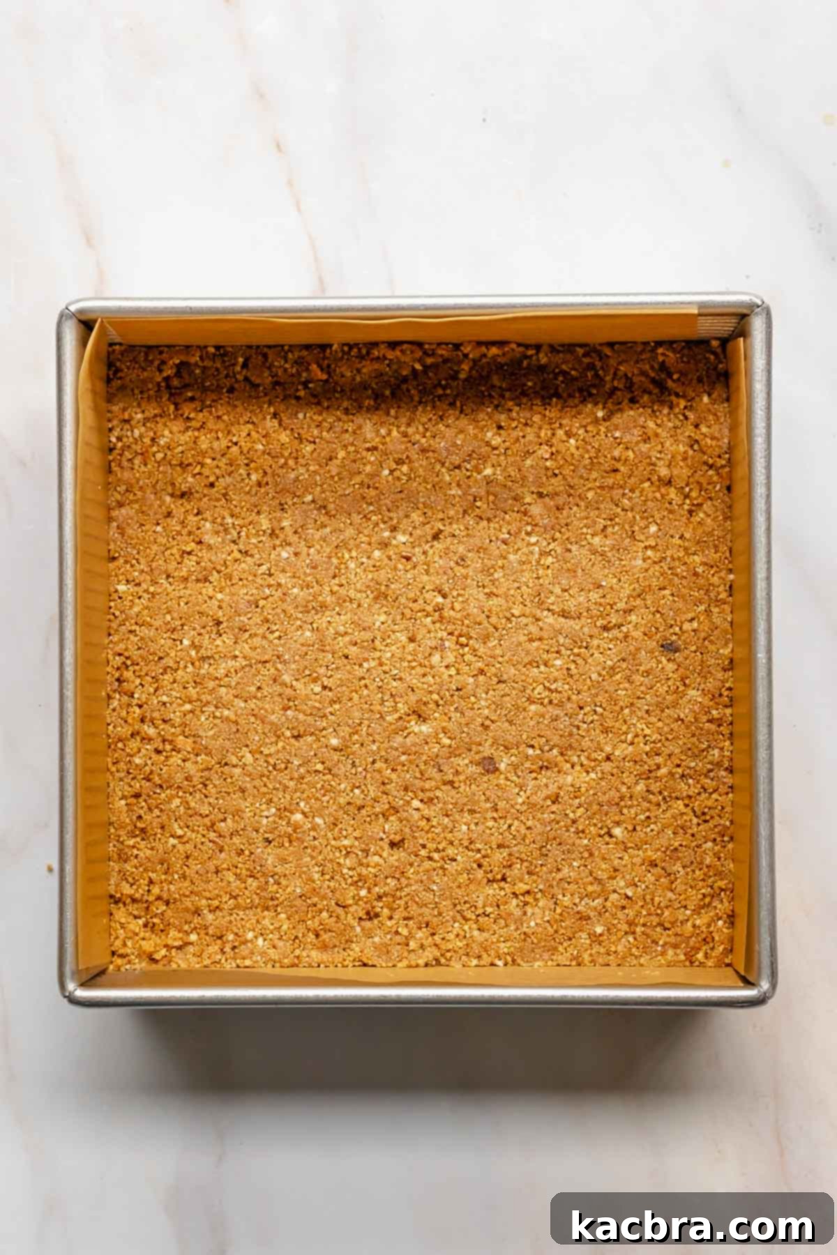 The graham cracker crust mixture firmly pressed into a square baking pan, ready for its initial bake.