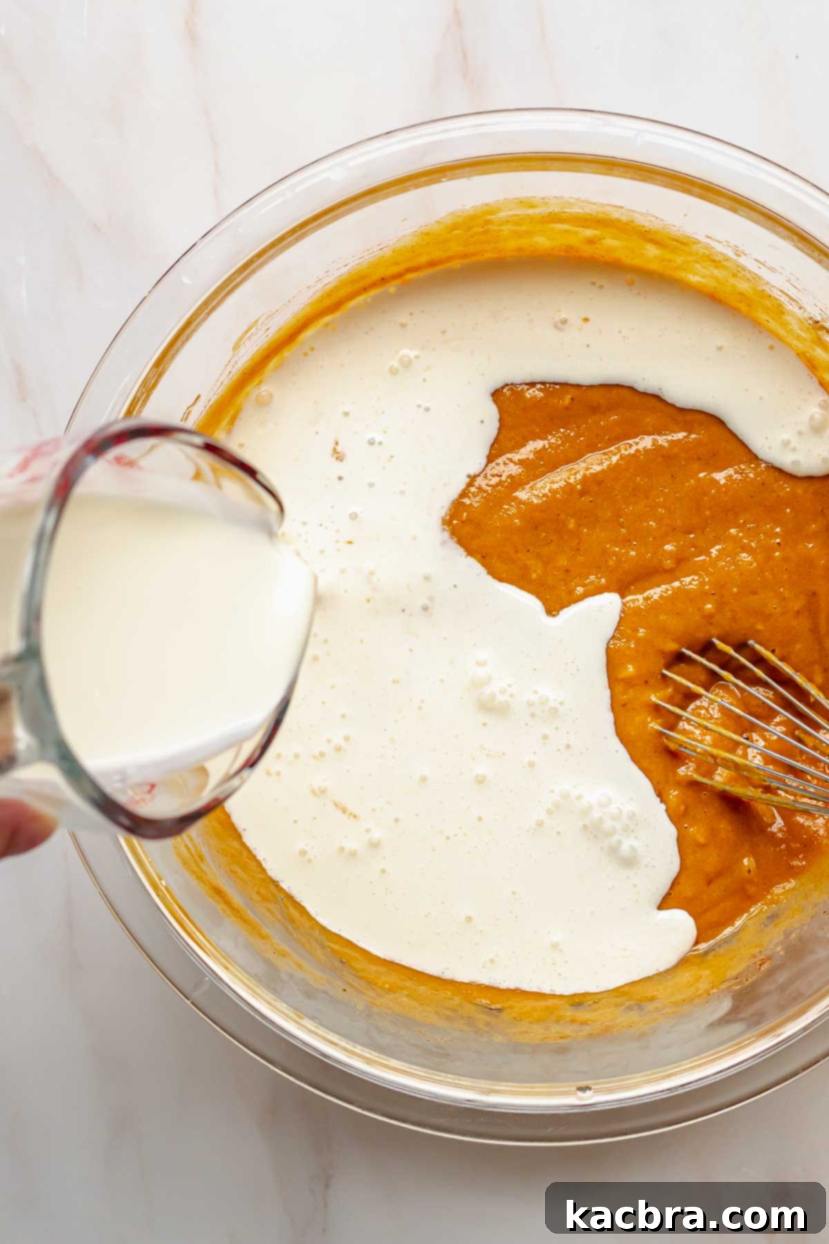 A hand gently pouring heavy cream into the sweet potato pie filling mixture.