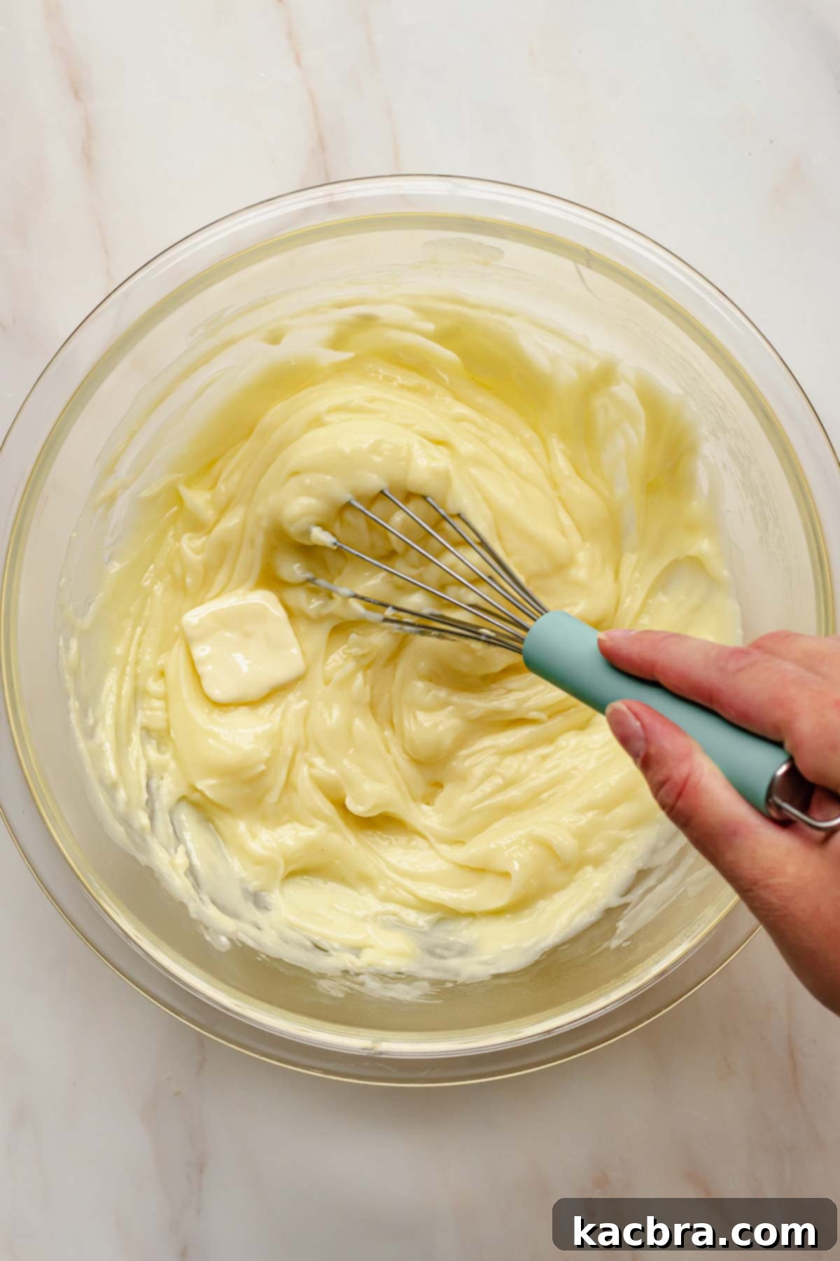 A hand whisking butter into warm vanilla pudding in a bowl.