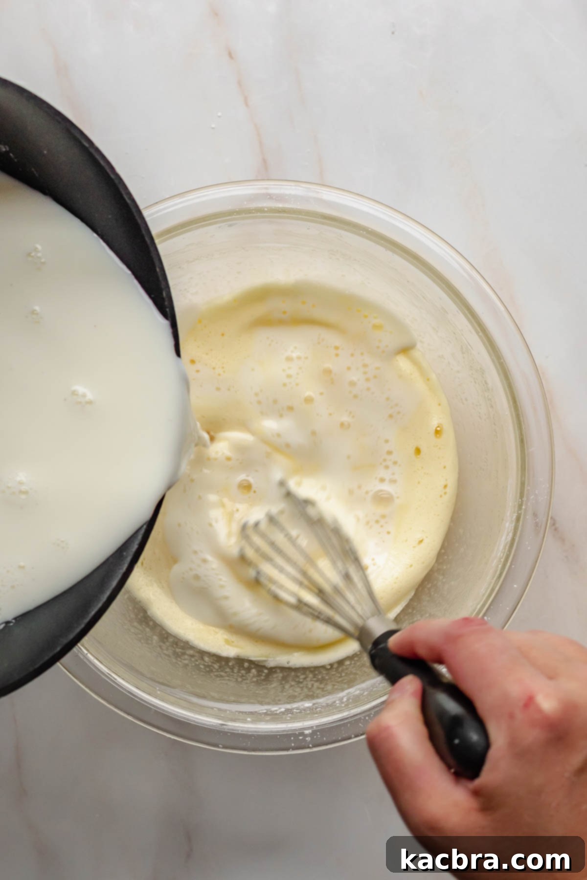 Hot milk mixture being slowly poured into the egg mixture while continuously whisking.