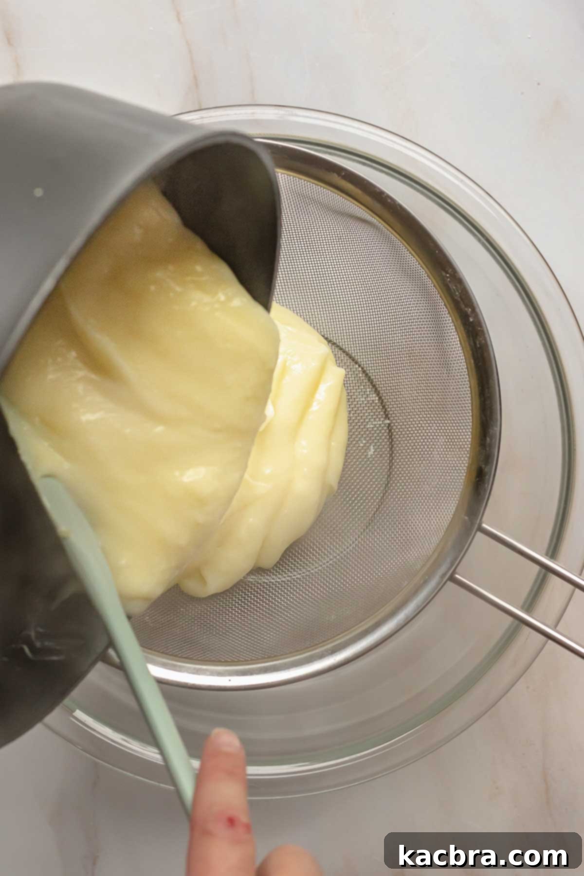 Thickened vanilla pudding being poured through a fine mesh sieve into a clean bowl.