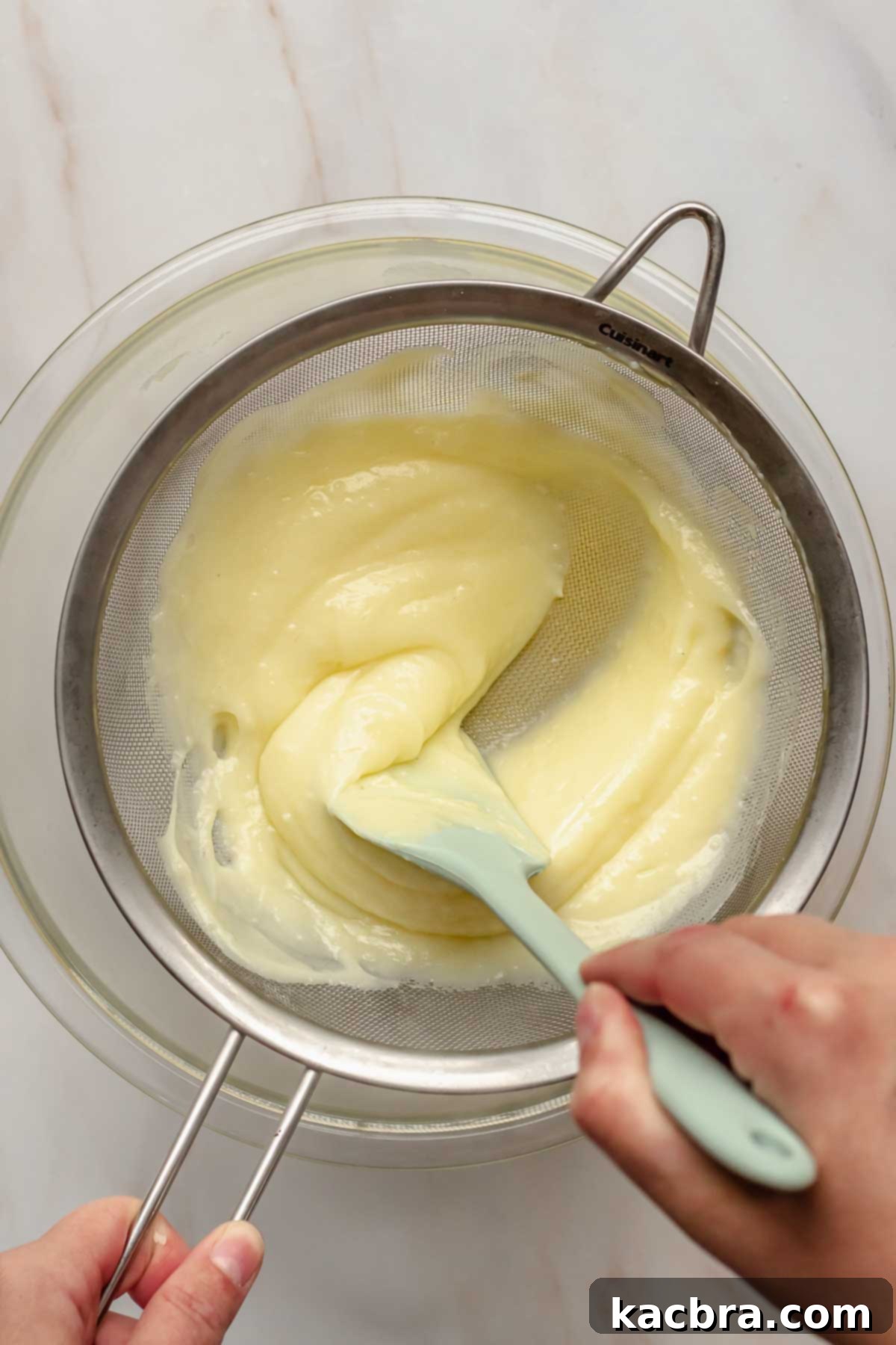 A rubber spatula pressing vanilla pudding through a fine mesh sieve to remove any lumps.
