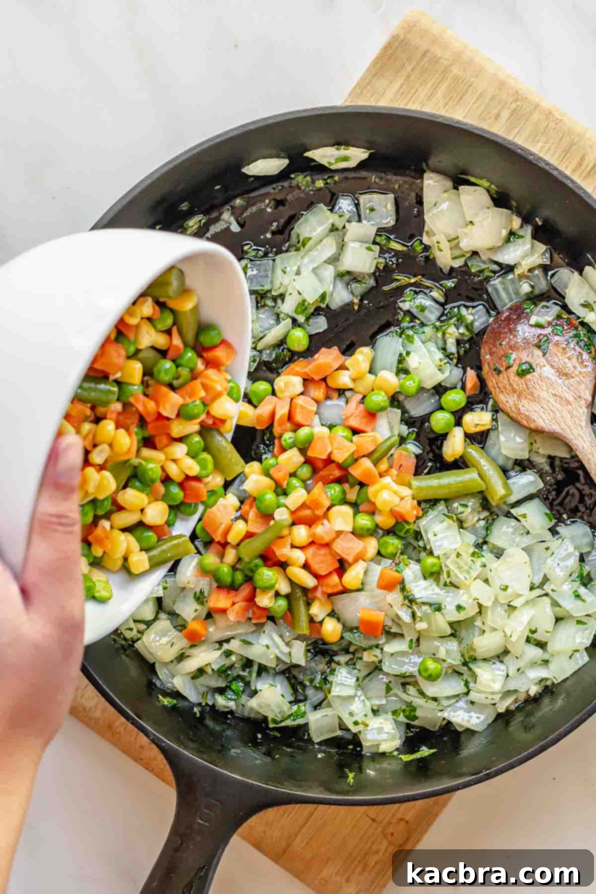 Mixed frozen vegetables being poured into a hot pan with sautéed onions and minced garlic, releasing steam.