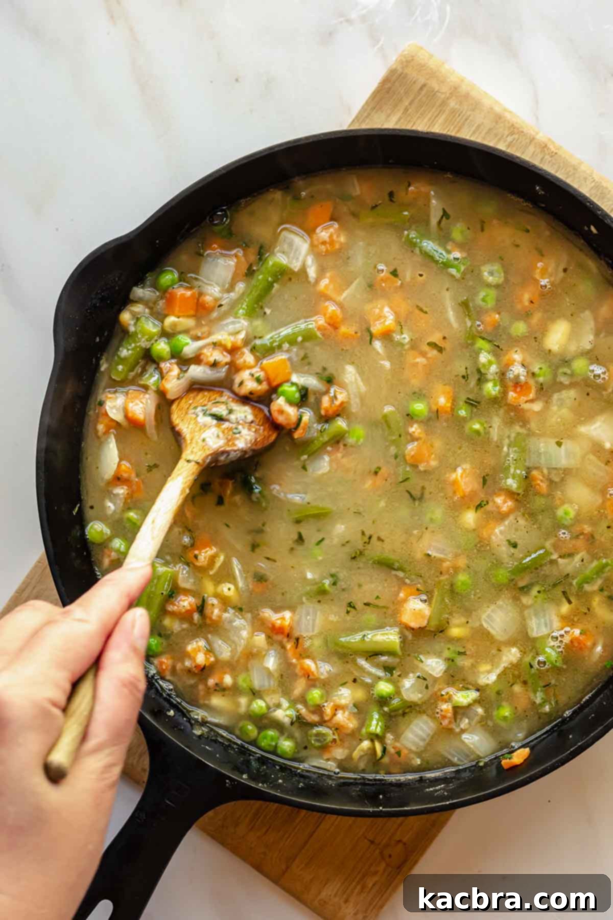 A wooden spoon stirs chicken stock into the vegetable and flour mixture in a pan, creating a smooth, bubbling gravy.