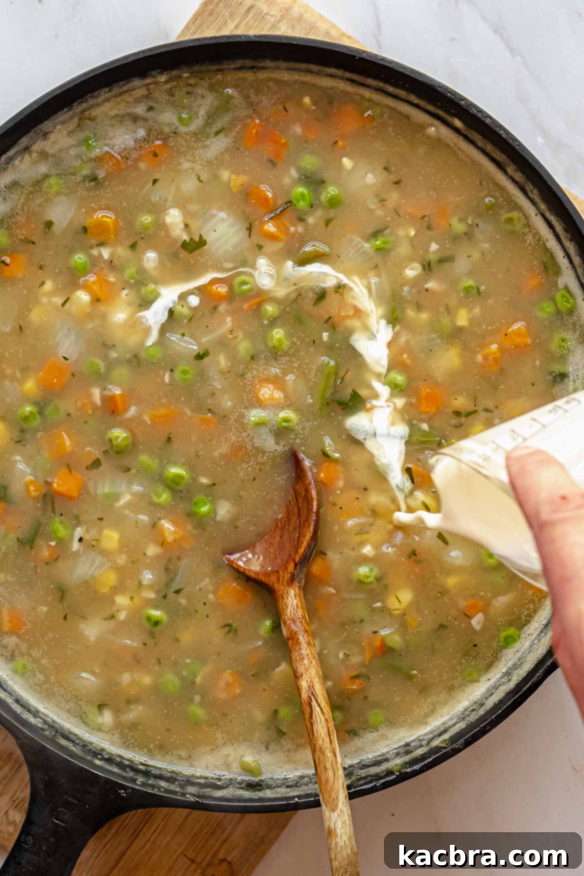A hand pouring heavy cream into the simmering pot pie filling in a pan, enriching the sauce.