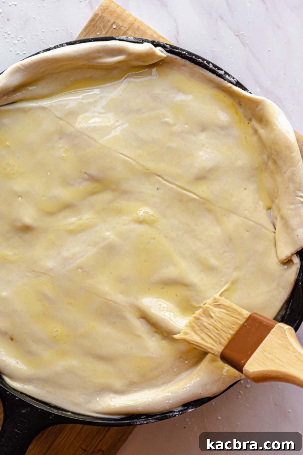 A pastry brush applying whisked egg wash over a sheet of puff pastry covering a pot pie, ensuring an even, golden coating.