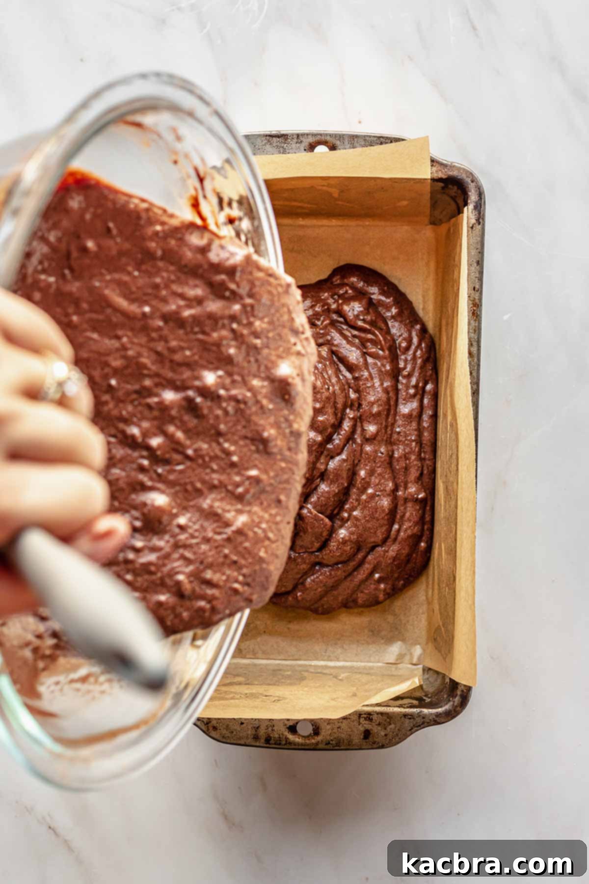 Decadent Chocolate Orange Bliss 10 A hand expertly pours the smooth chocolate cake batter from a mixing bowl into a parchment-lined loaf pan, preparing it for baking.