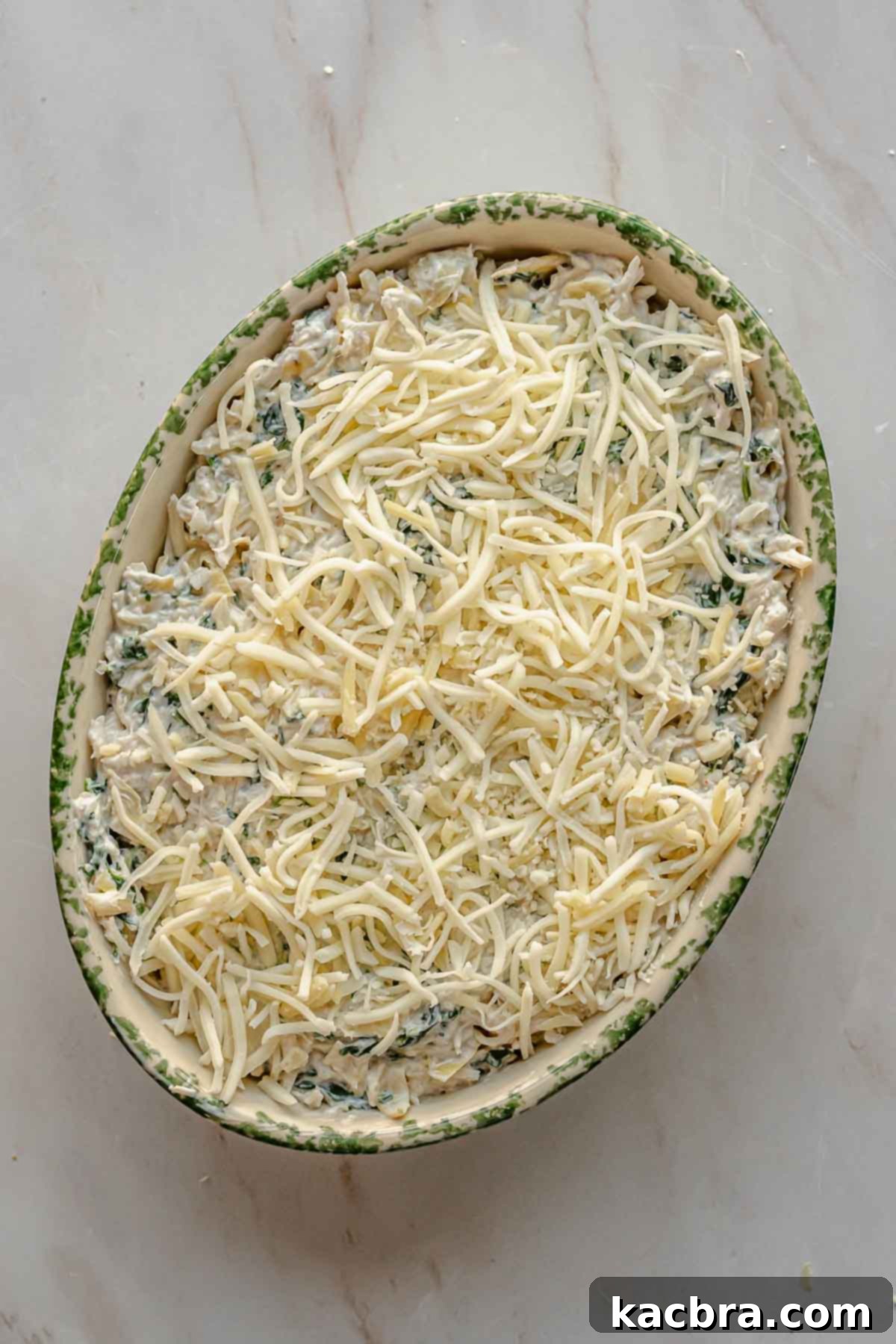 Shredded cheese is sprinkled over the unbaked crab spinach artichoke dip in a casserole dish.