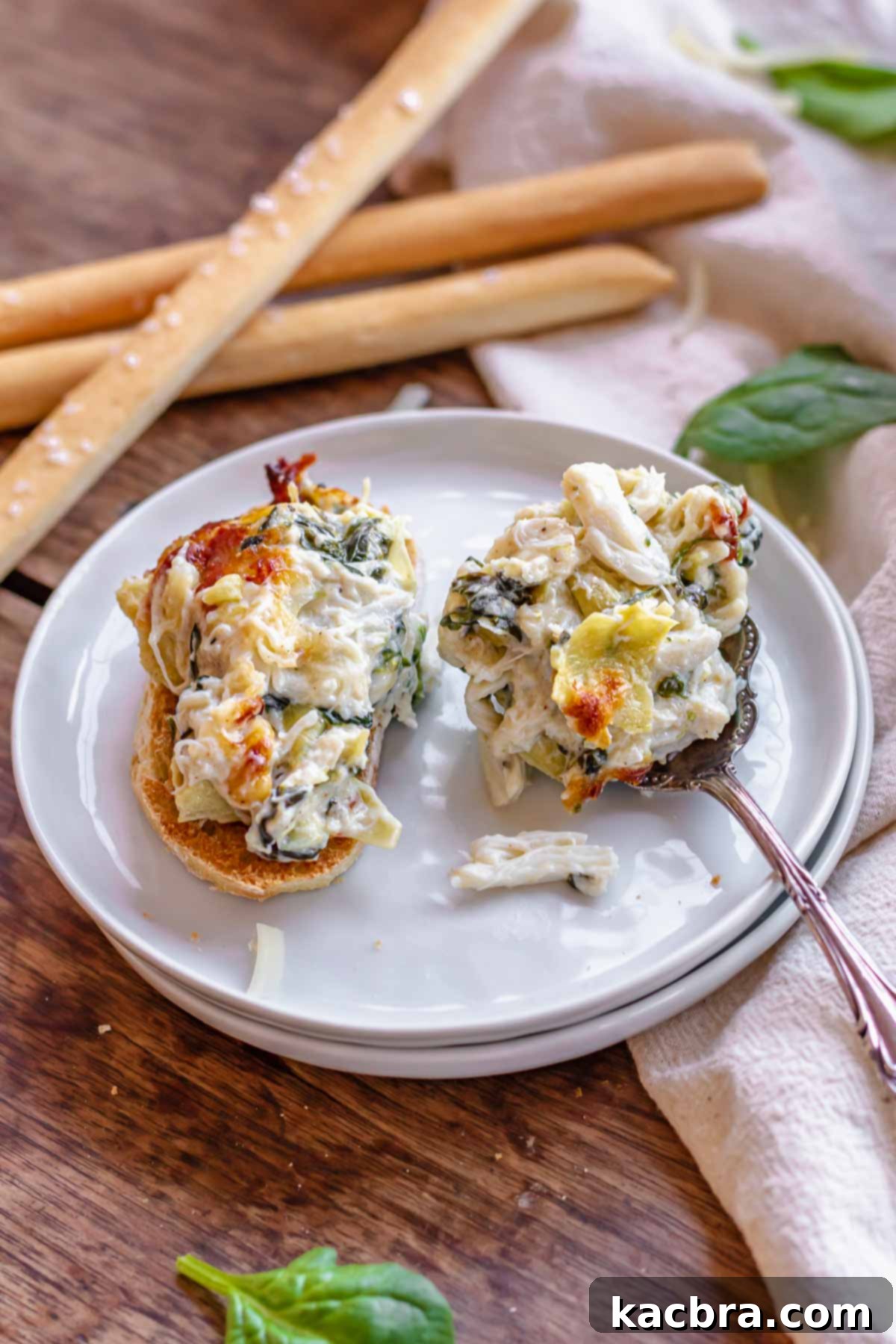A spoonful of spinach artichoke crab dip on a toasted baguette slice, with more dip on a spoon resting on a plate.