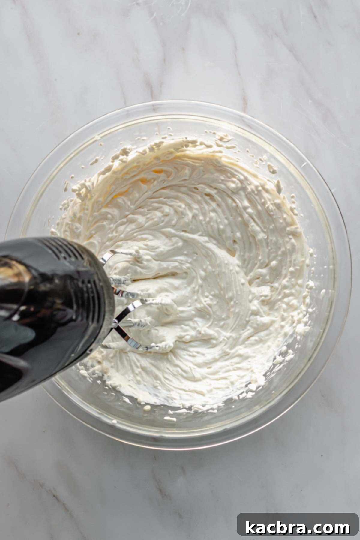 A hand mixer blends cream cheese, mayonnaise, and sour cream in a bowl until smooth and fluffy.