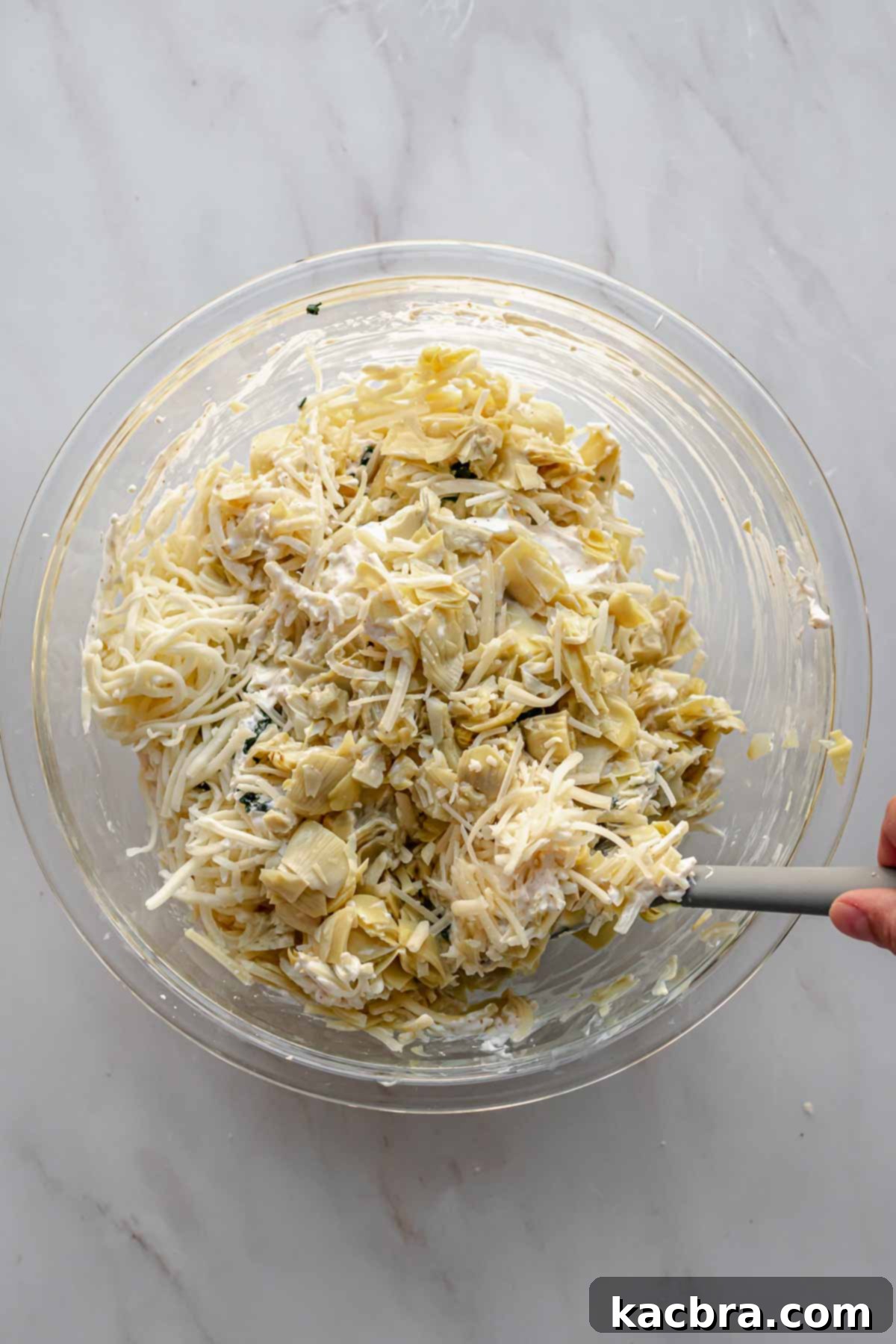 A spatula is used to mix the spinach, artichokes, and cheeses into the creamy dip base.