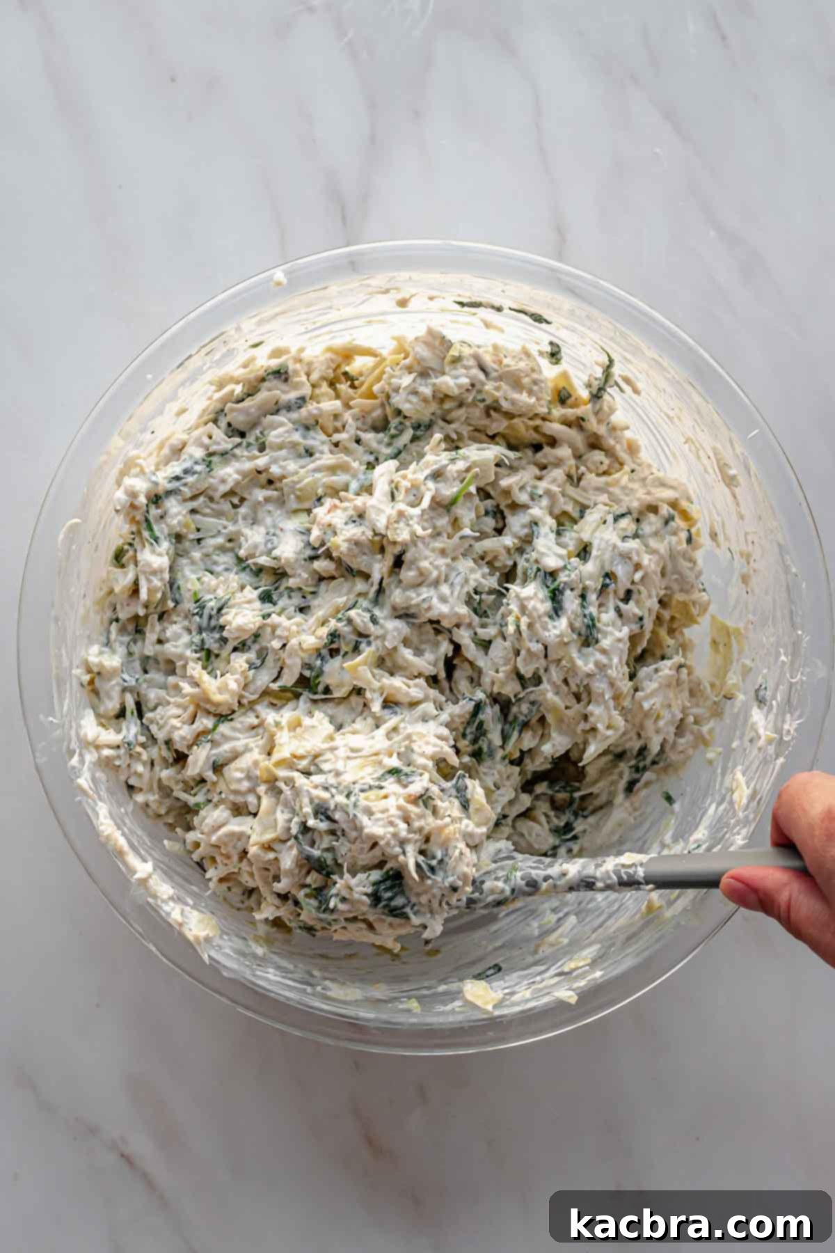 A rubber spatula gently folds crab meat into the spinach artichoke dip to avoid breaking up the lumps.