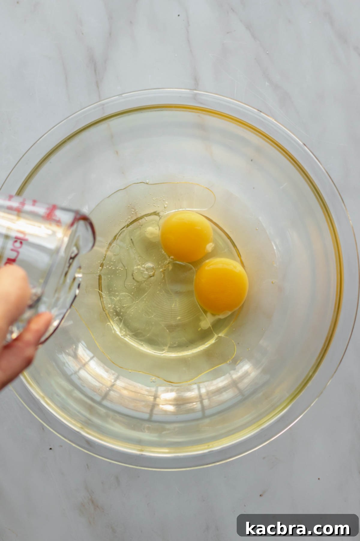 A glass measuring cup pours oil into a bowl with eggs.