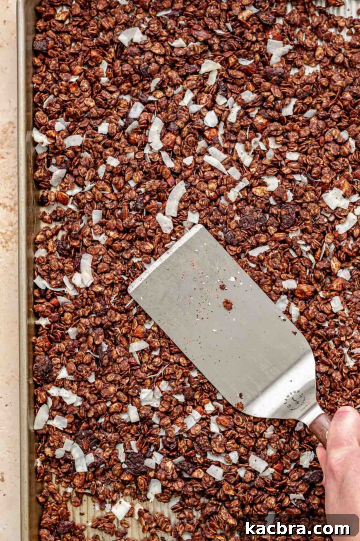 A spatula pressing down granola on a sheet pan after adding coconut flakes.