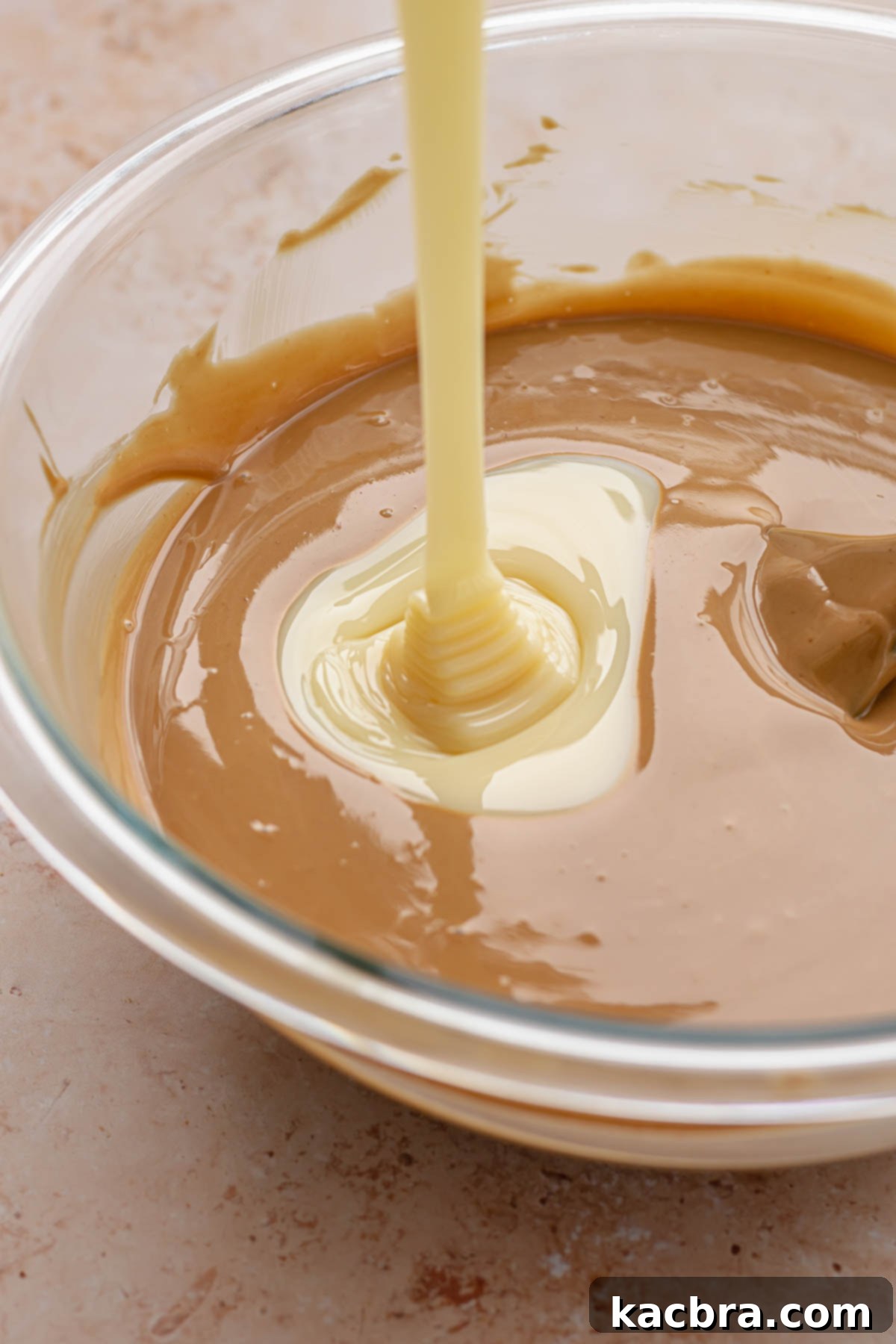 Sweetened condensed milk pouring into a bowl of melted white chocolate and Biscoff spread.