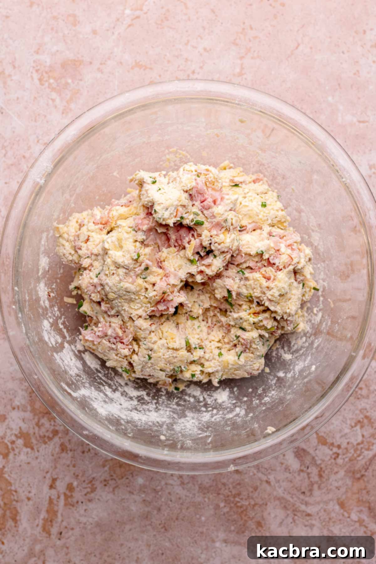 The finished ham and cheese scone dough, mounded beautifully in a bowl. The image captures the dough's slightly shaggy texture and the visible pieces of ham, cheese, and chives.